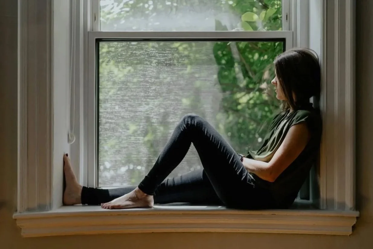 Woman sitting thoughtfully on a window sill looking outside.