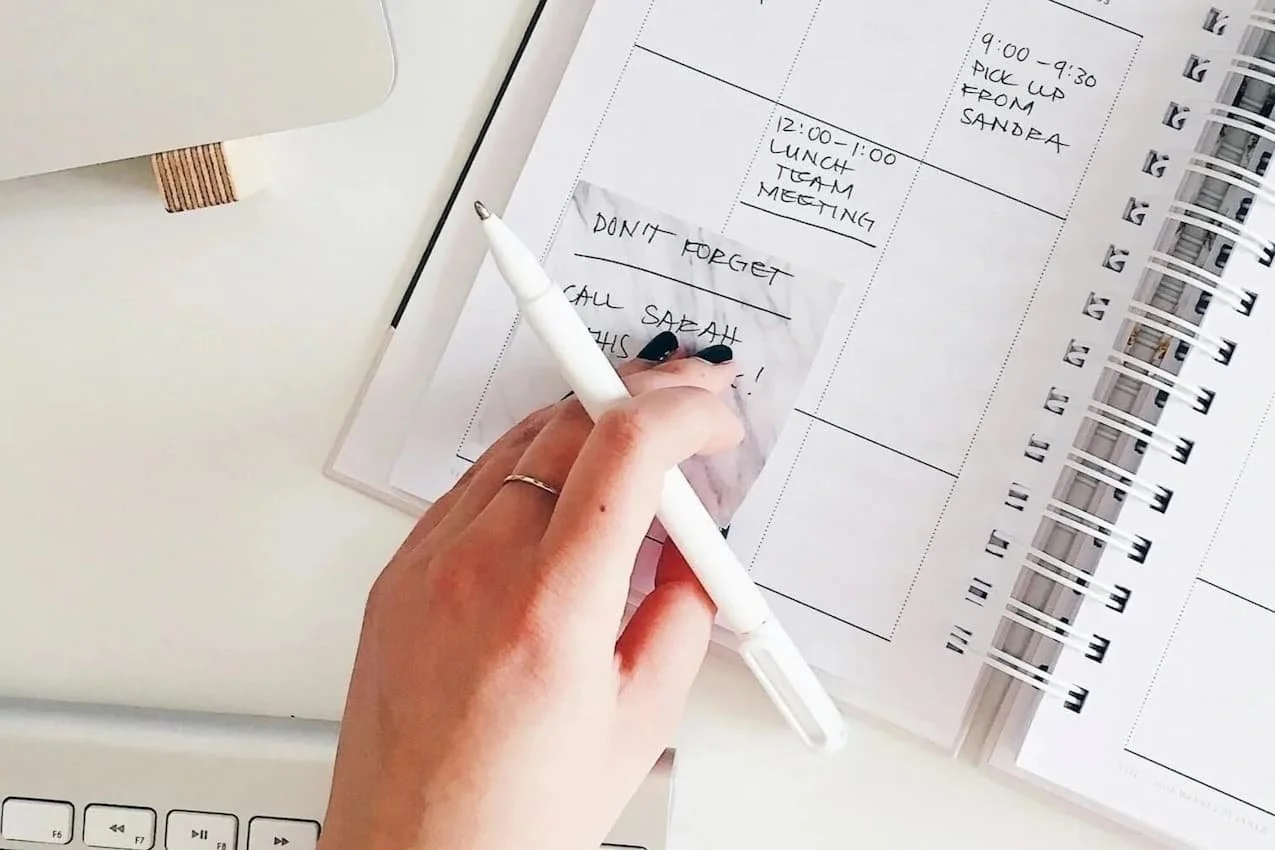 A hand holds a pen over an open weekly planner, writing on a sticky note that reads 'Don't forget, call Sarah,' next to a keyboard and laptop.