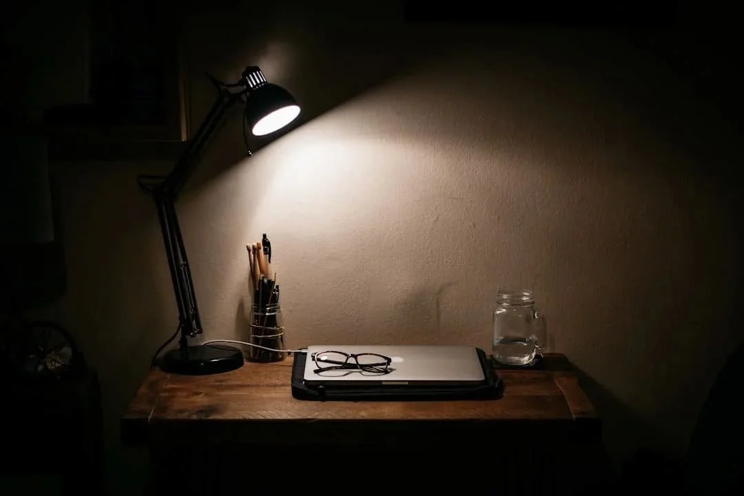 Desk lamp glowing in a dark room, illuminating glasses and a jar on a wooden table