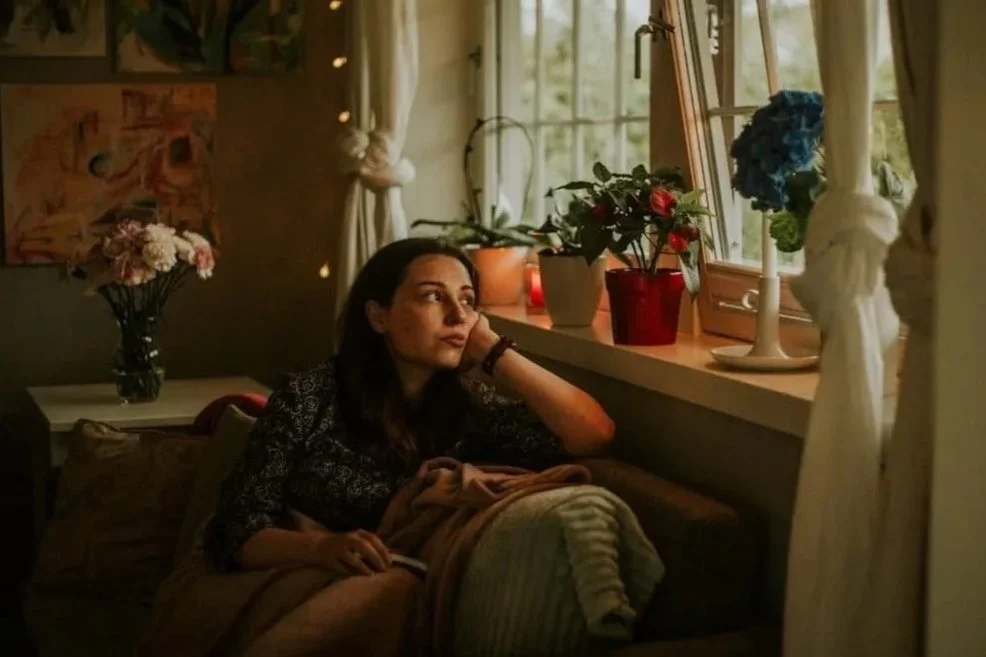 A woman rests her chin on her hand, gazing thoughtfully out a window surrounded by plants in a warmly lit room.