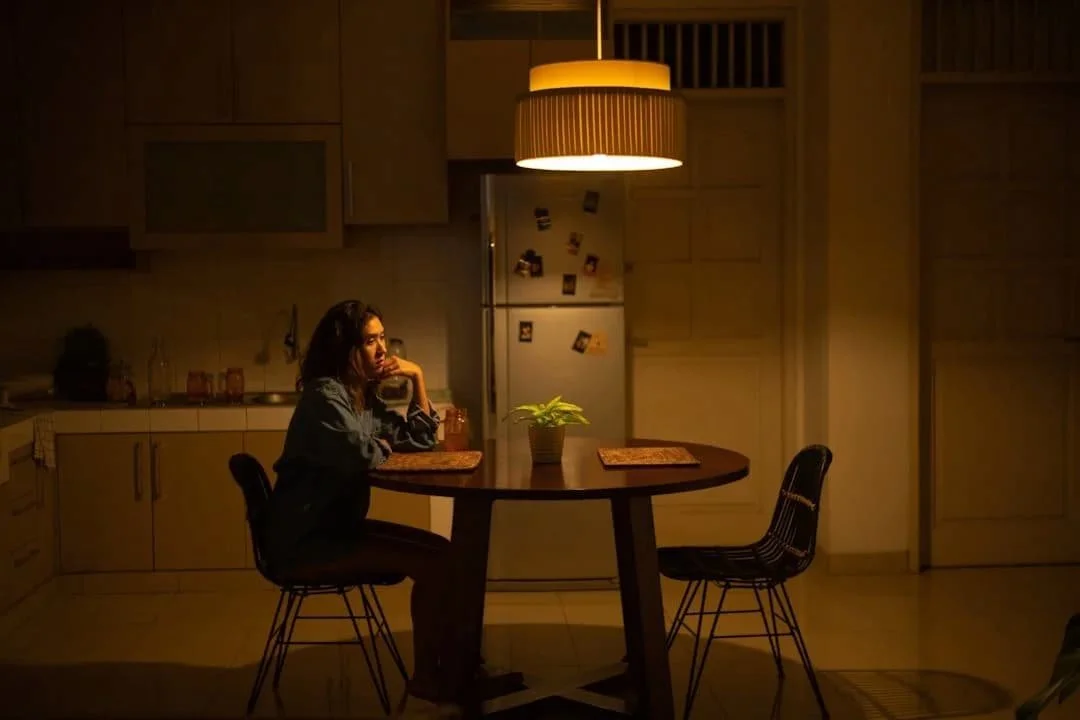 A woman sits alone at a kitchen table in the evening, resting her chin on her hand.