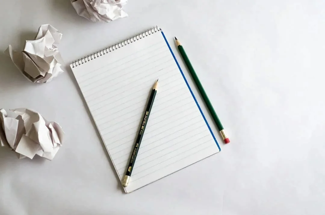 A blank spiral notepad with two pencils on a white surface, surrounded by several crumpled sheets of paper.