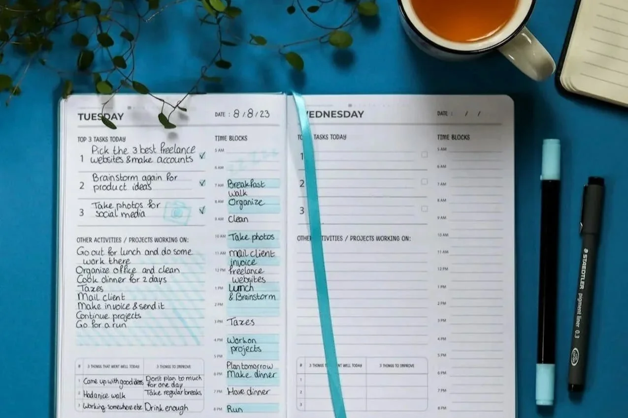 An open weekly planner with completed tasks on one page and a blank page beside it, next to tea and pens.