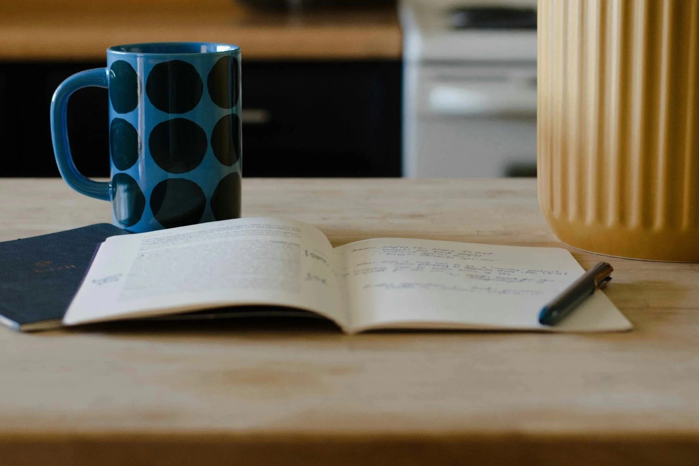 A blue polka-dot mug beside an open notebook with handwritten notes and a pen on a wooden kitchen counter.