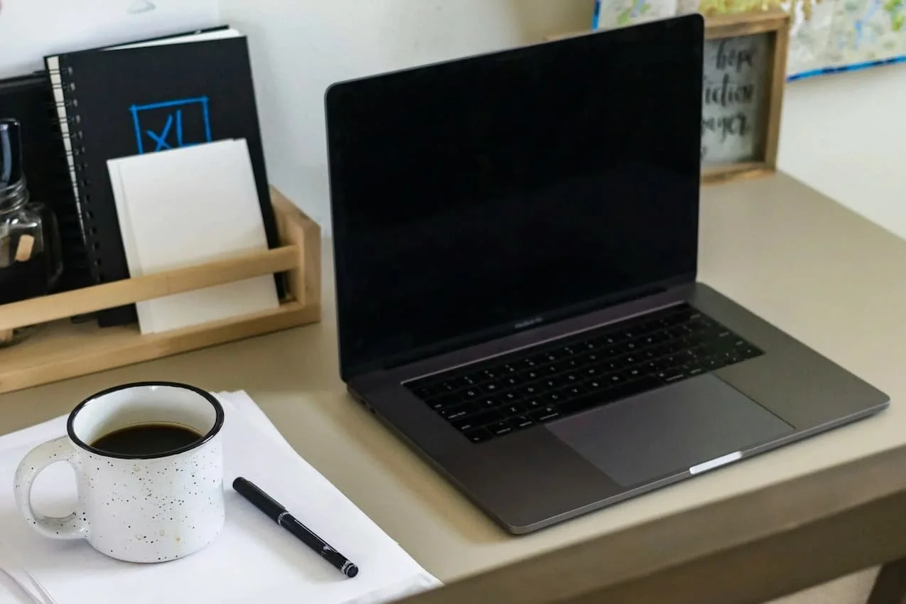 Open laptop on a home office desk beside a coffee mug, pen, and stack of papers, suggesting work waiting to be started.