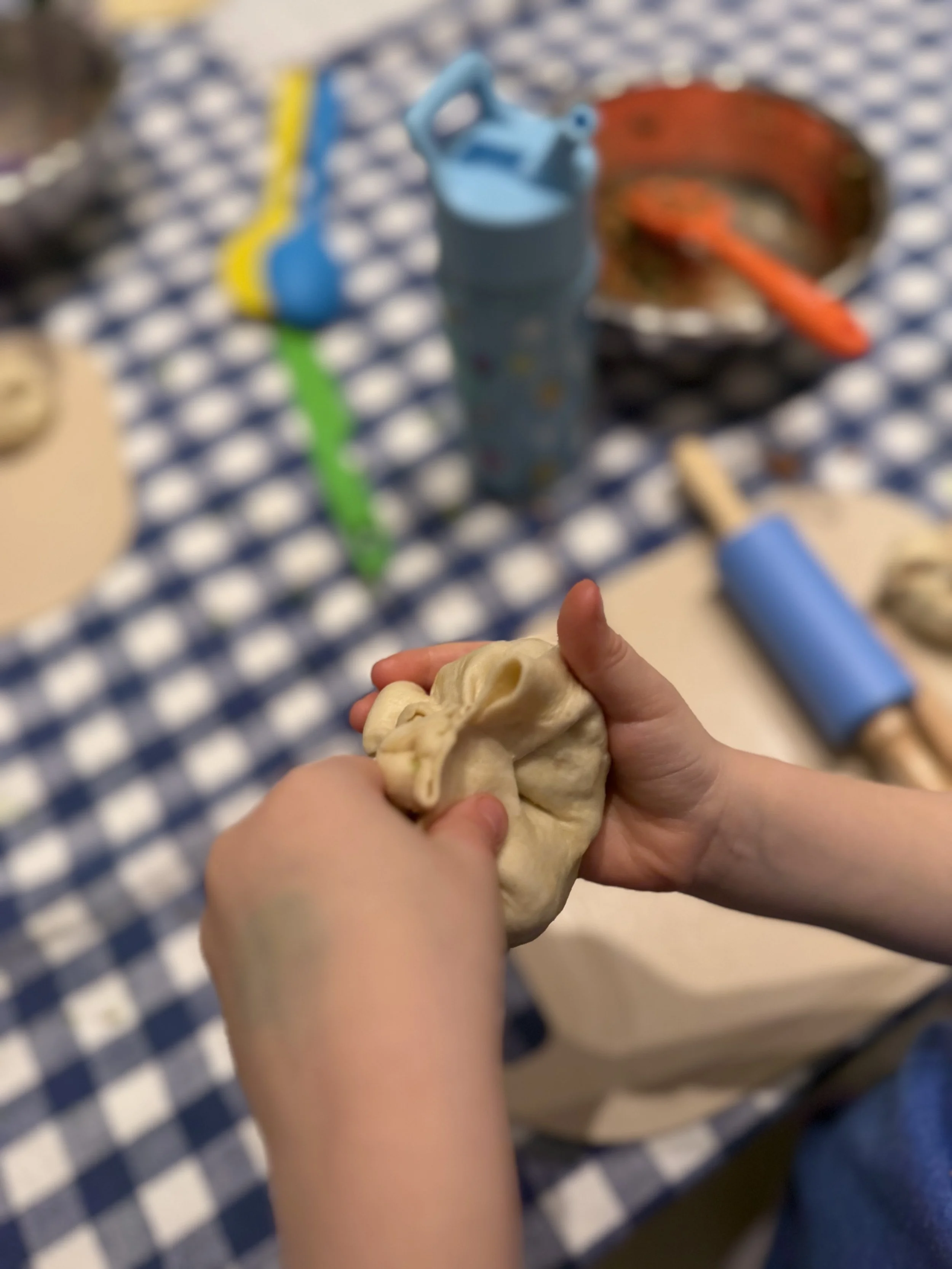 Child folding and shaping raw pizza dough on a checkered blue and white tablecloth.
