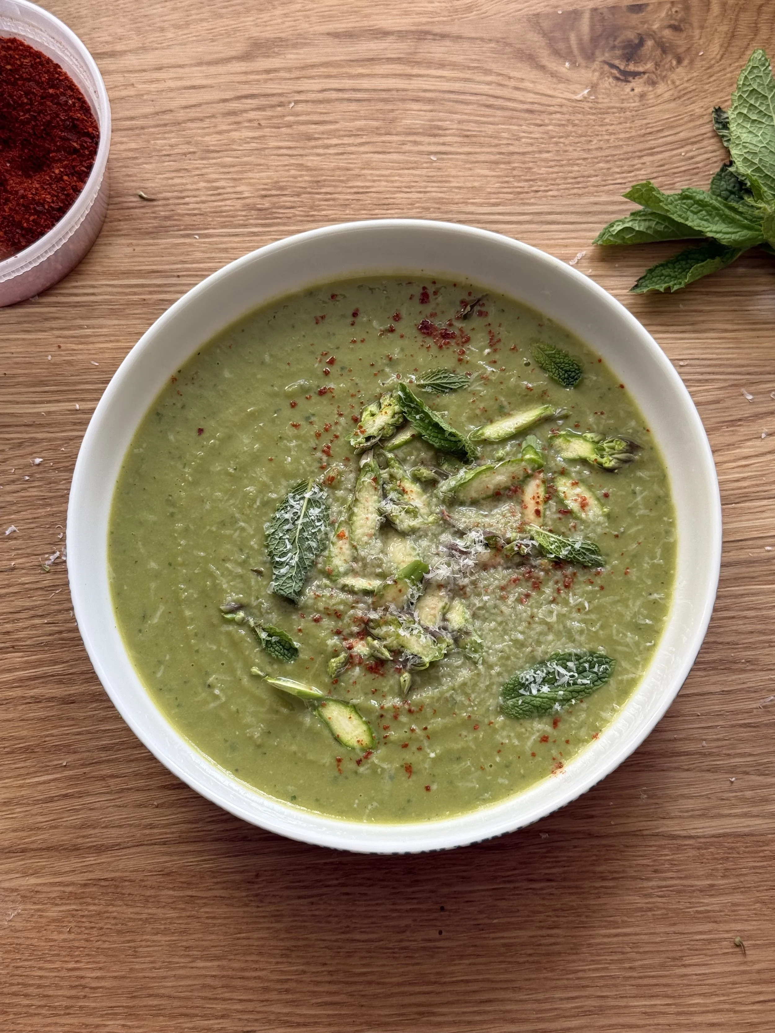 Bowl of green vegetable soup garnished with herbs and spices, placed on a wooden surface with fresh mint leaves and a container of chili powder nearby.