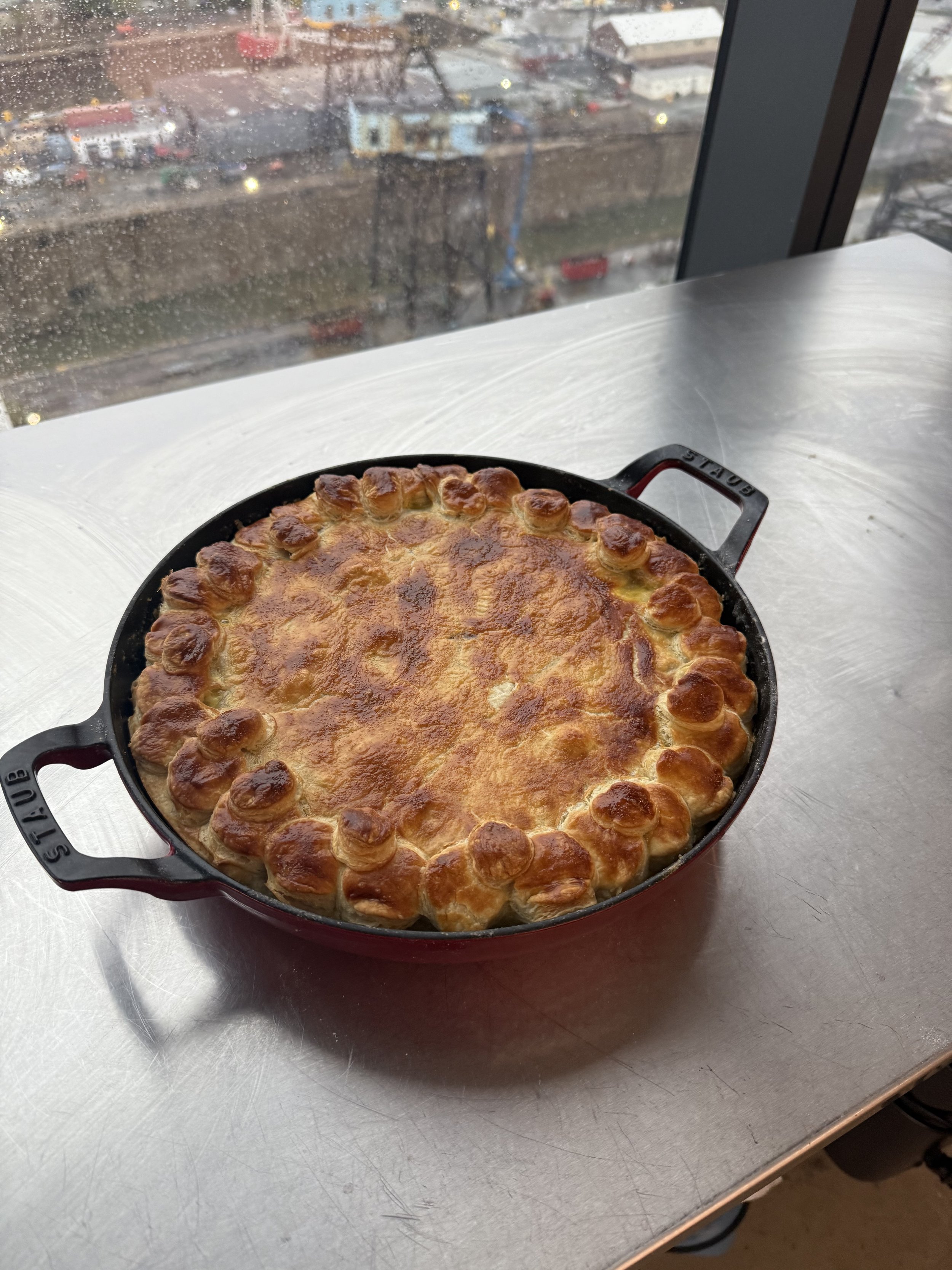 Baked dish with browned cheese crust in a black cast iron skillet on a white table near a window with rainy weather outside.