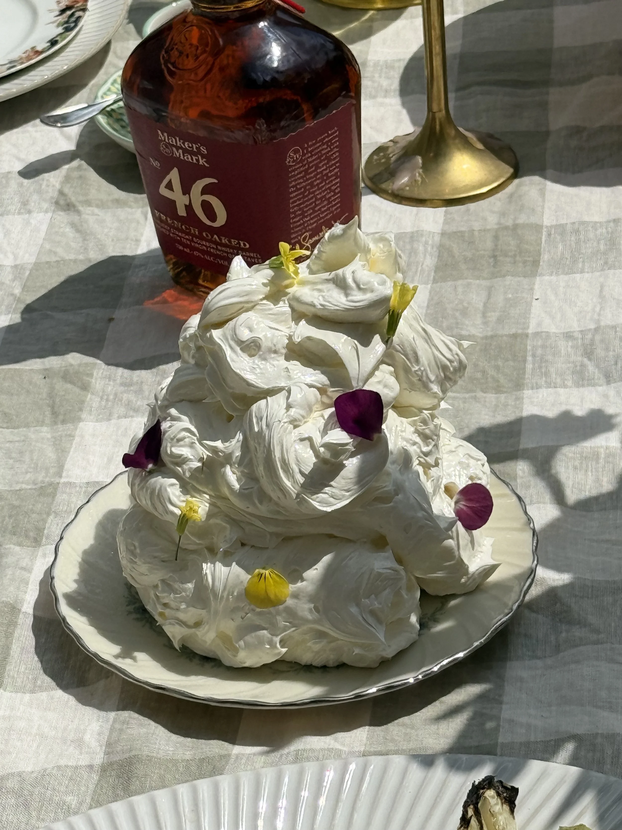 A plate of meringue with edible flower petals on top, sitting on a checkered tablecloth, with a bottle of Maker's Mark bourbon and a gold candlestick in the background.