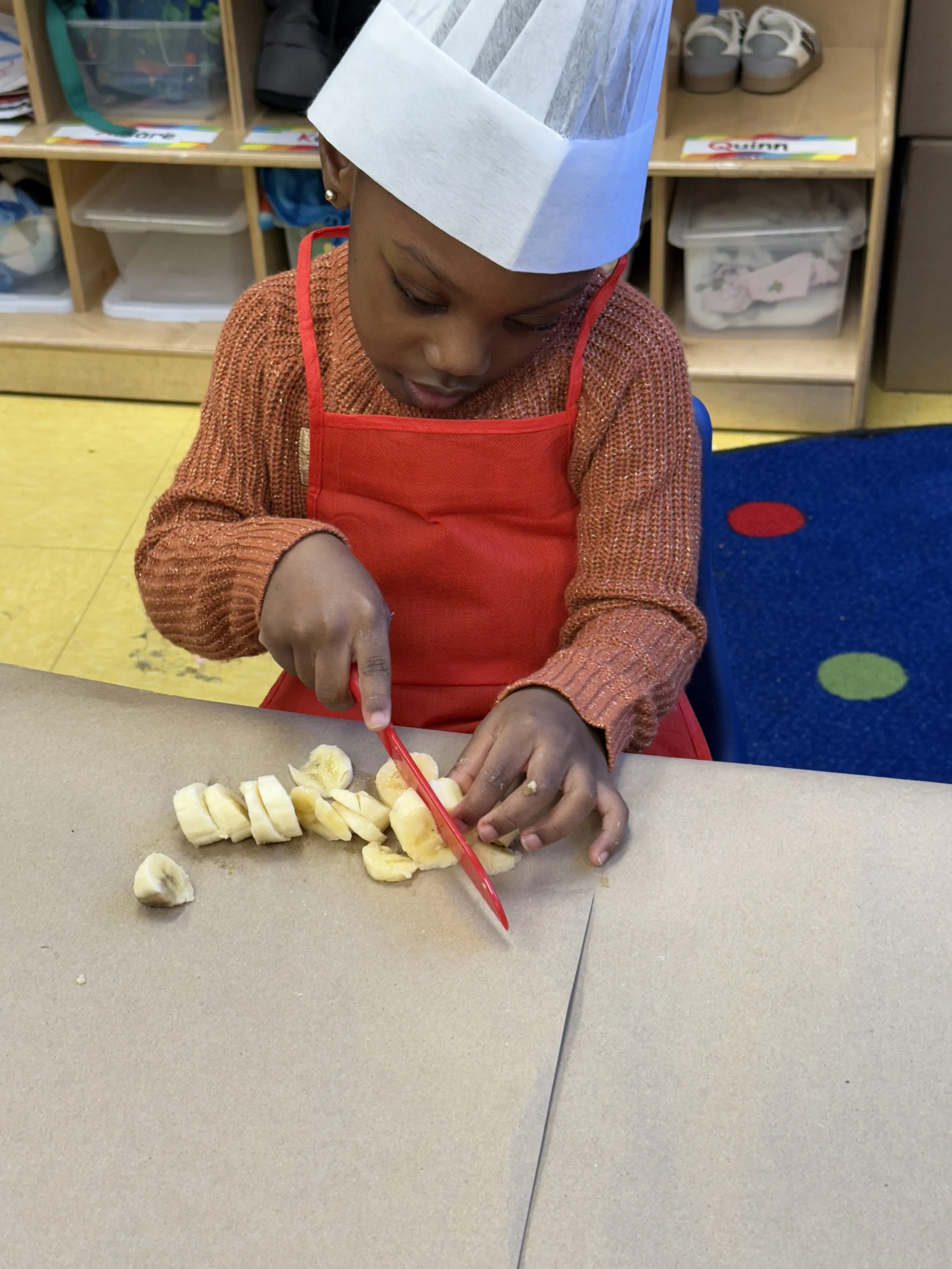 A young child wearing a paper hat and red apron cutting bananas with a red plastic knife at a classroom table.