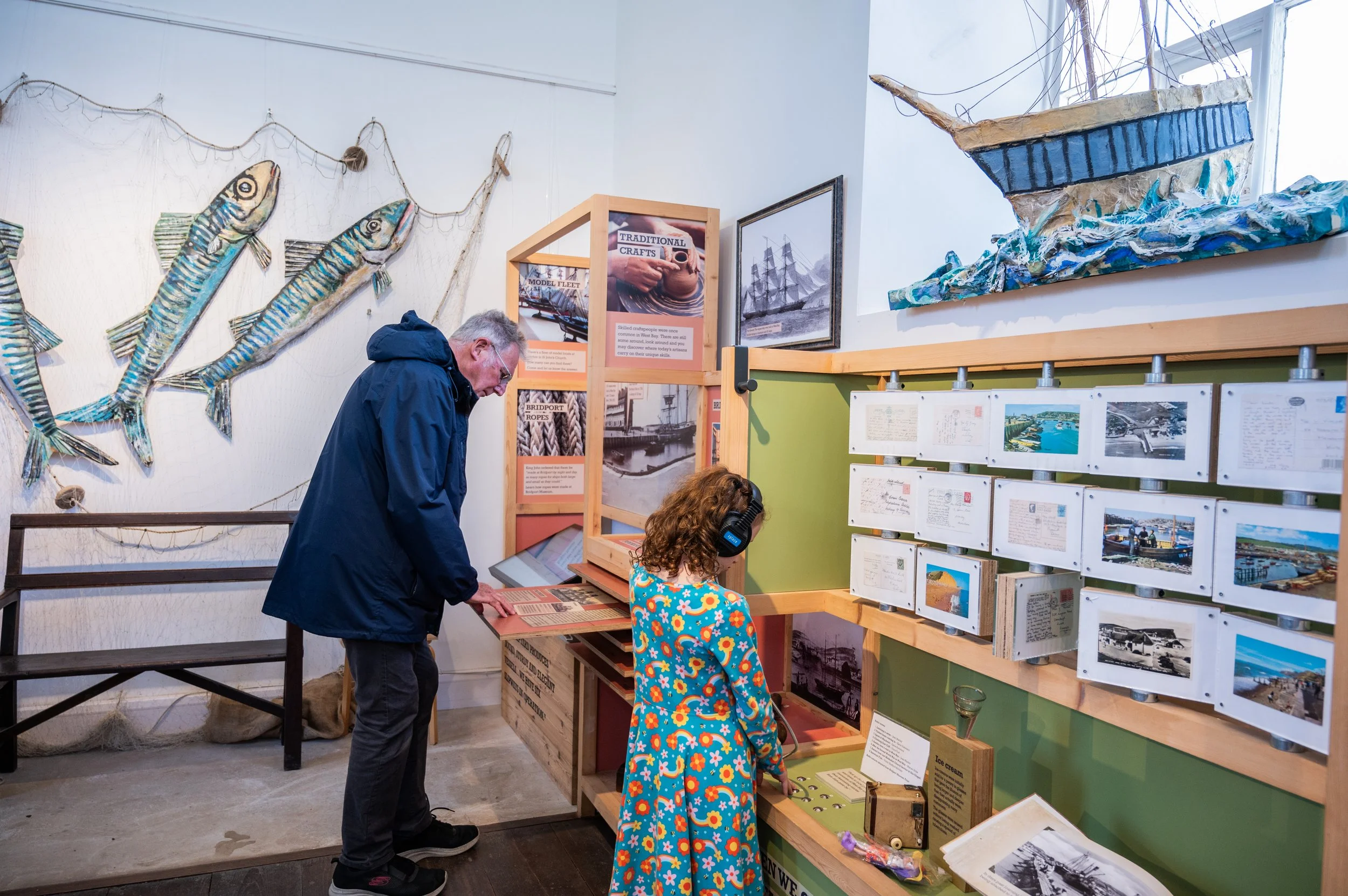 an older man pulling out a wooden board to find out more information and a young girl facing the display wearing headphones
