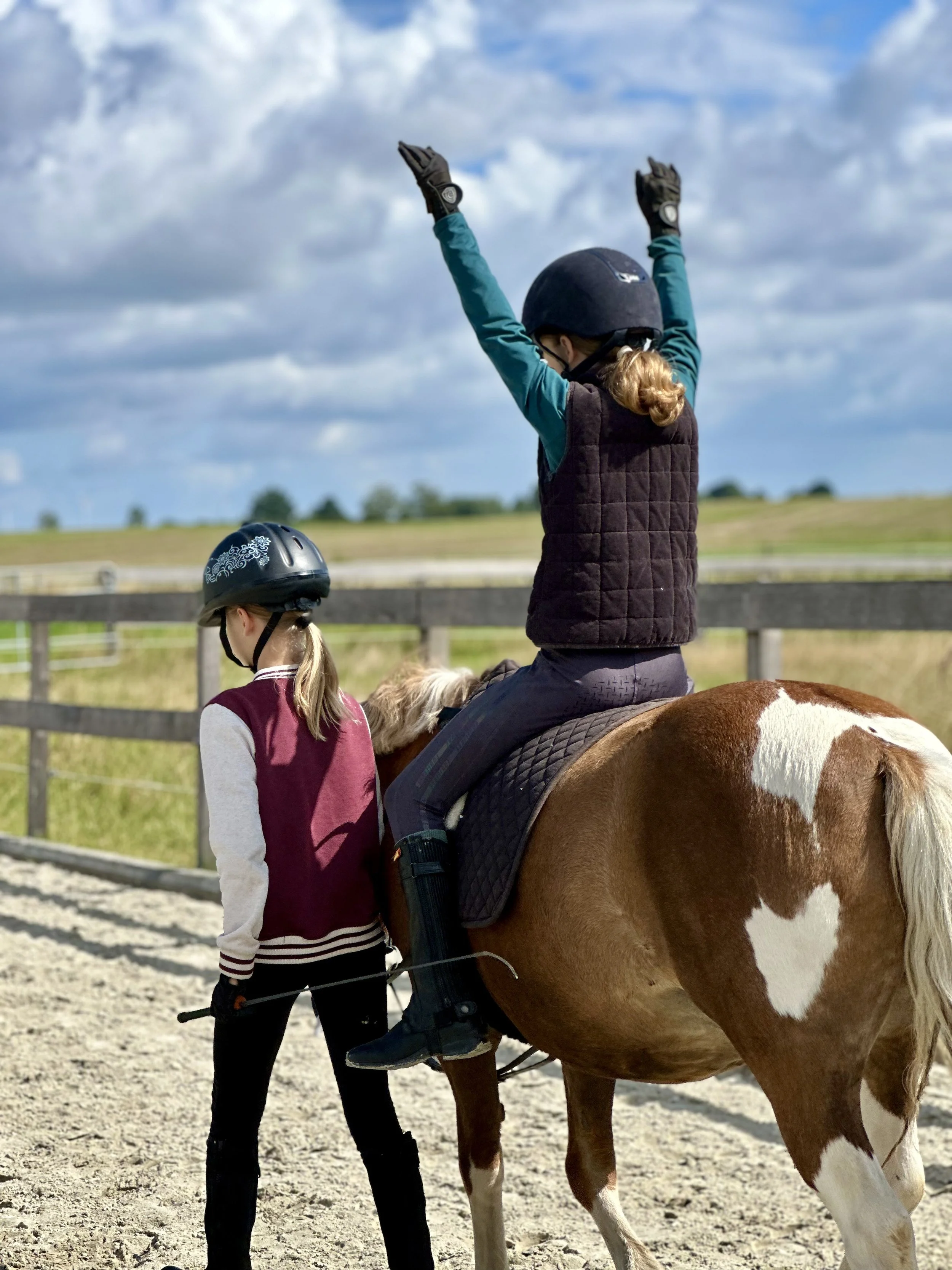 Ein Kind führt ein Pony während ein anderes Kind auf dem Pony sitzt und die Arme ausstreckt.