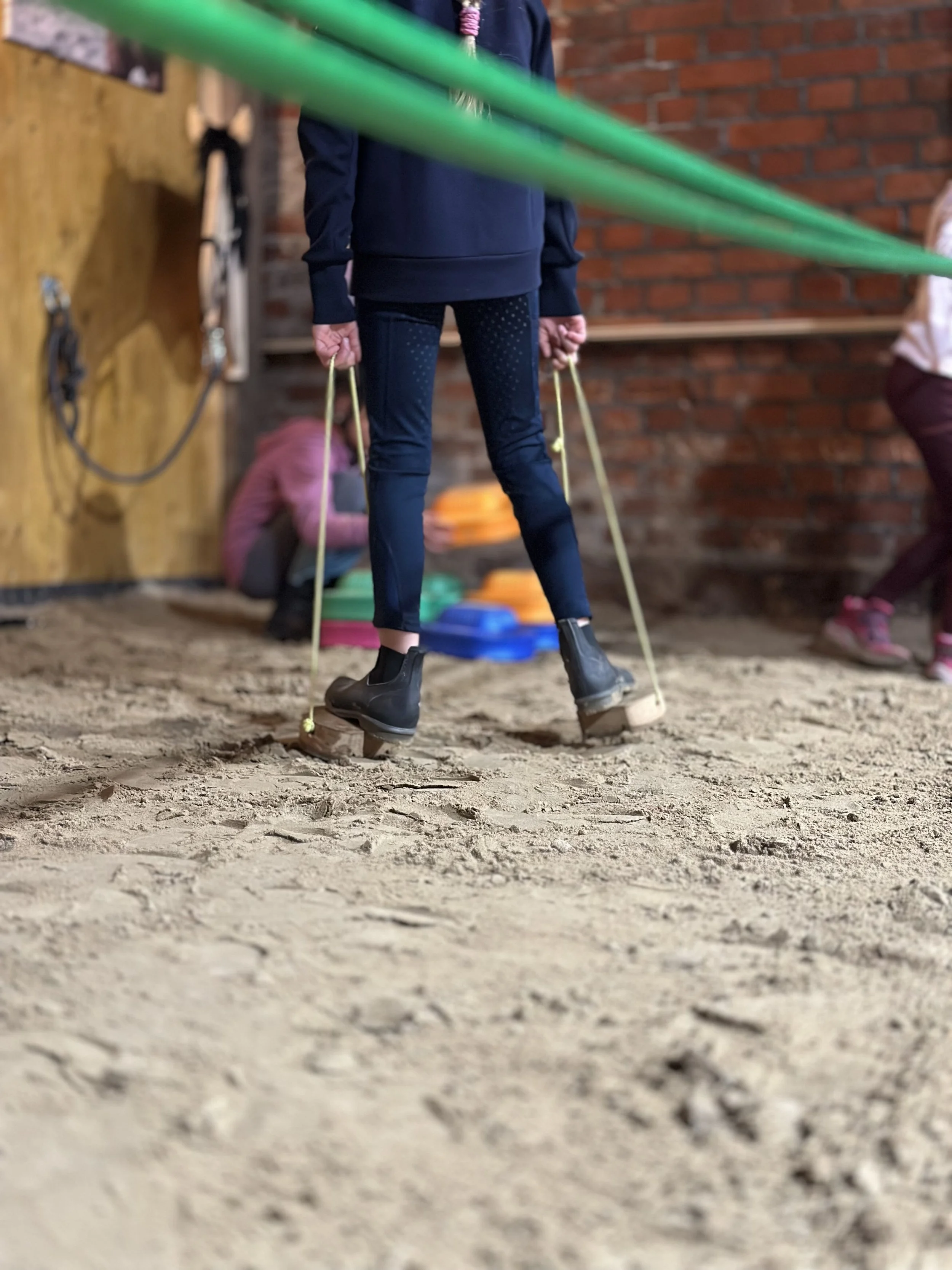 Ein Kind in schwarzen Stiefeln und dunkelblauen Hosen läuft mit Huf-Stelzen auf Sand mit anderen Kindern im Hintergrund.