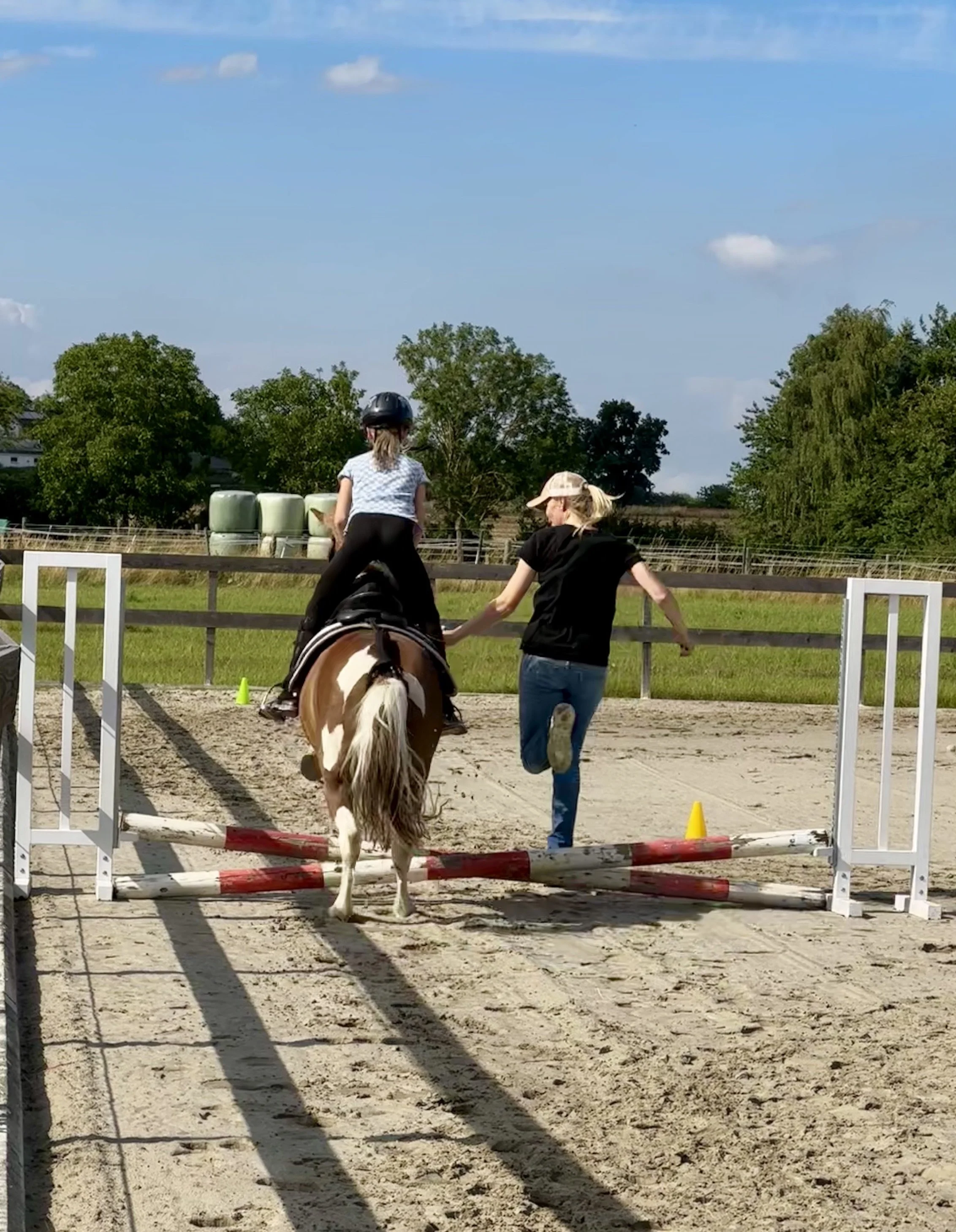 Ein junges Mädchen reitet auf einem Pony über einen kleinen Sprung auf dem Reitplatz im Freien, begleitet von einer Frau, die neben ihr her läuft und sie begleitet.