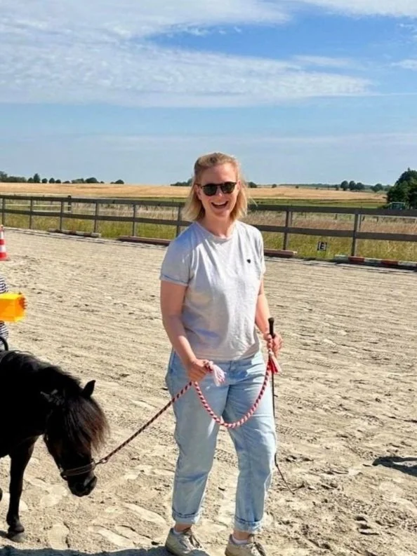 Eine lächelnde Frau mit Sonnenbrille steht auf einem sandigen Reitplatz und hält einen Strick an einem kleinen schwarzen Pferd oder Pony, mit offenen Feldern und einem blauen Himmel mit einigen Wolken im Hintergrund.