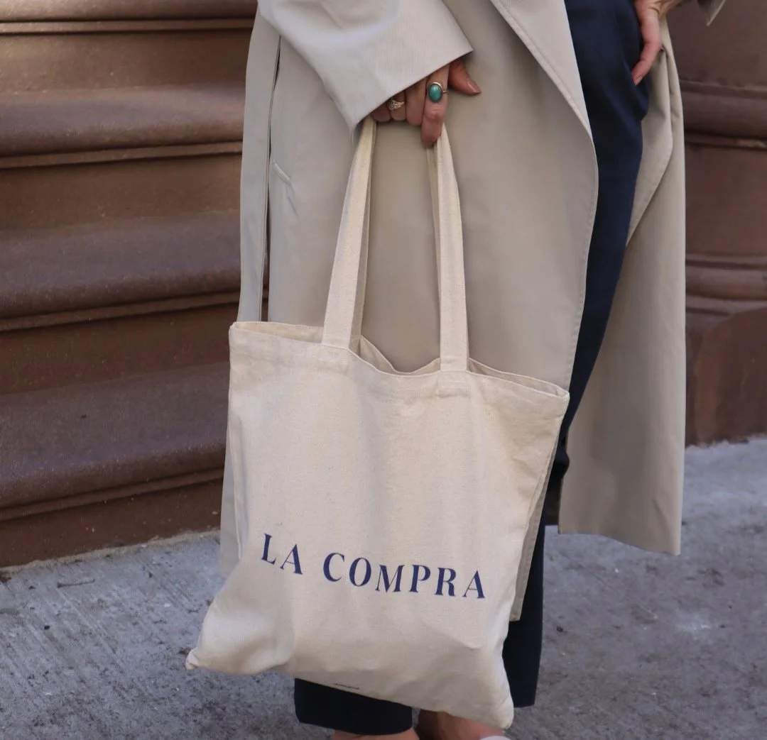 A person wearing a beige coat holding a beige tote bag with the words 'LA COMPRA' printed in blue, standing on a sidewalk near brown stairs.