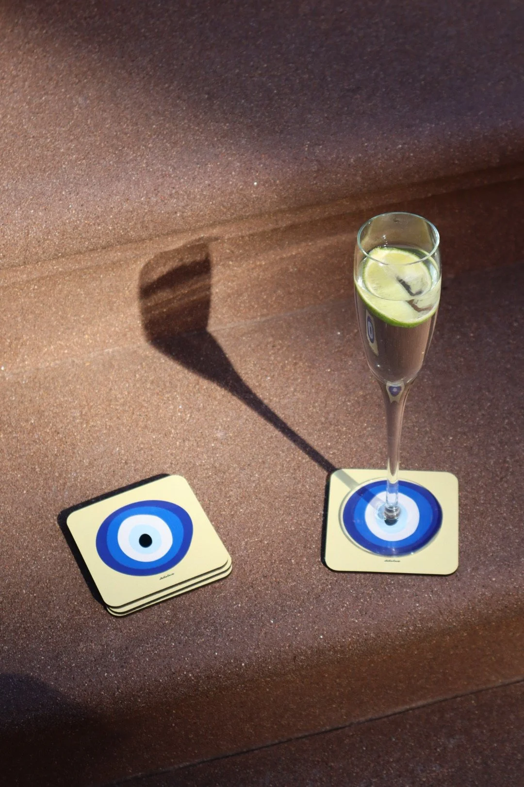 A glass of sparkling water with lime slices, casting a shadow on a textured red surface. Next to the glass are several coasters with blue concentric circles on a light yellow background.