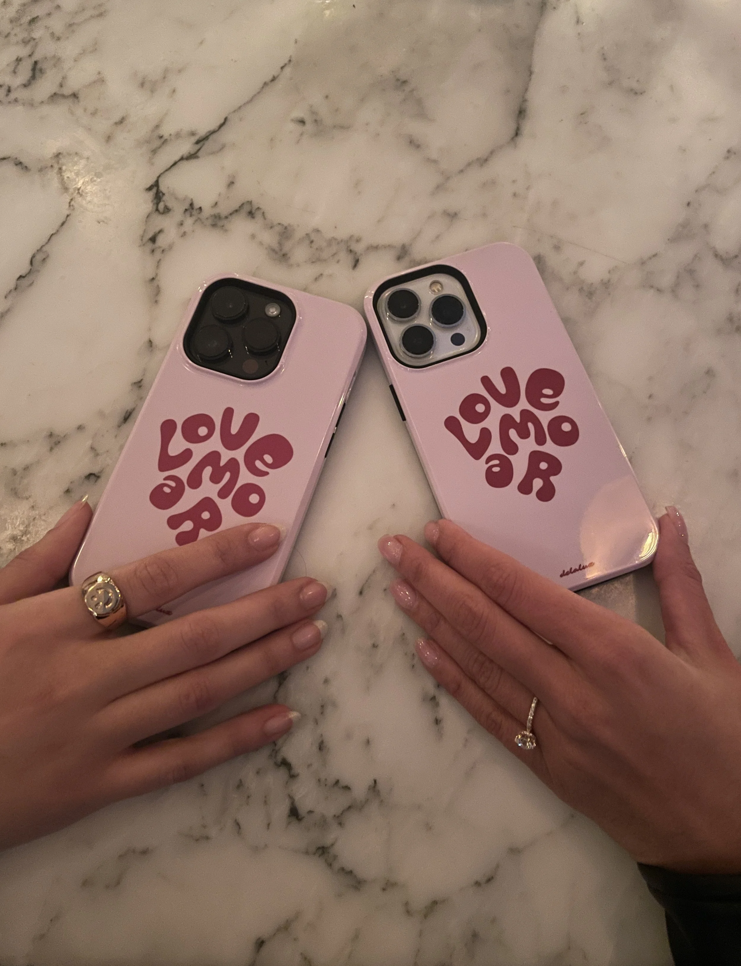 Two hands with rings holding pink phone cases with red cartoon text that says 'LOVE MORE' on a marble surface.