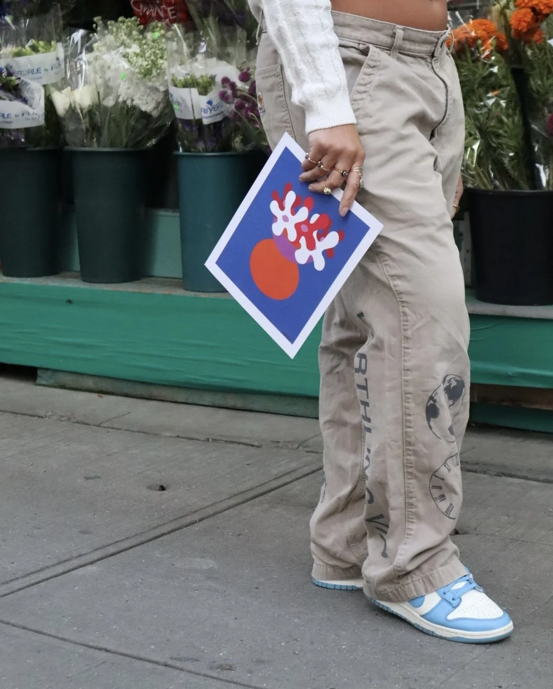 Person standing outside a floral shop holding a colorful greeting card with abstract shapes and snowflakes, wearing beige pants, white sneakers with blue accents, and rings on fingers.