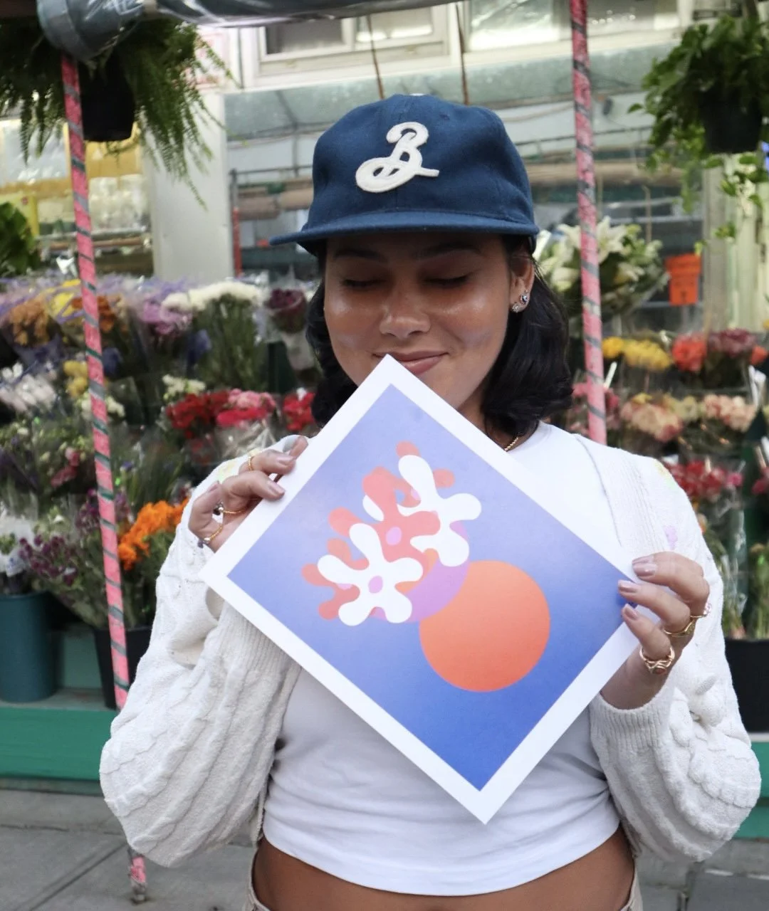 Young woman with a blue baseball cap and earrings, smiling with closed eyes, holding an abstract art print depicting coral and a round shape, in a flower market.