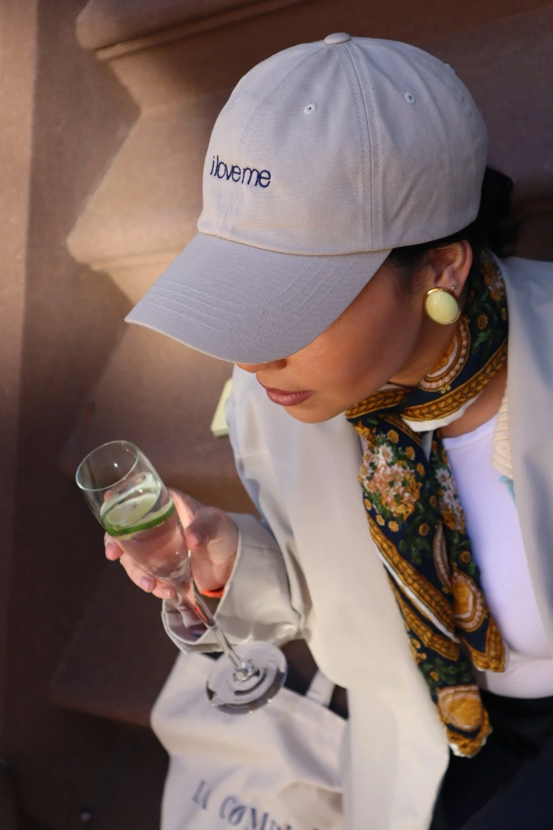 A woman wearing a gray baseball cap that says 'lovenme,' large round earrings, a patterned scarf, and a white jacket, holding a glass of sparkling water with lime.