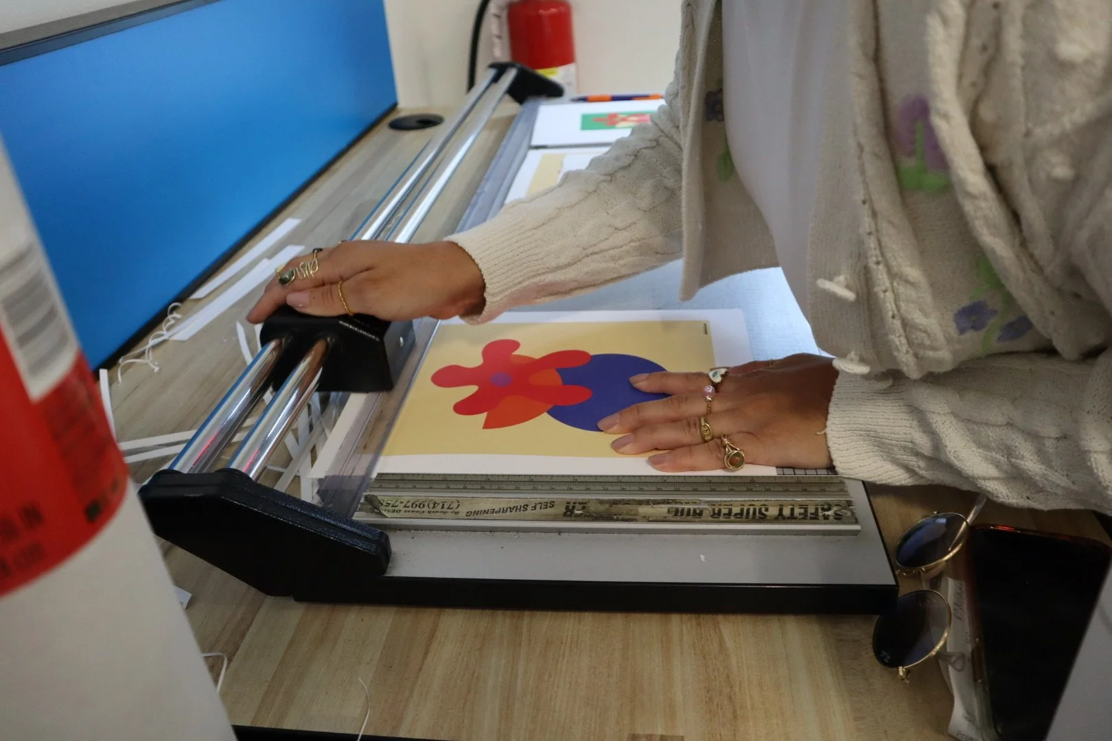 Person using a paper cutter to trim a colorful abstract artwork on paper, wearing rings and a cream-colored cable-knit sweater, with sunglasses and a phone on the table nearby.