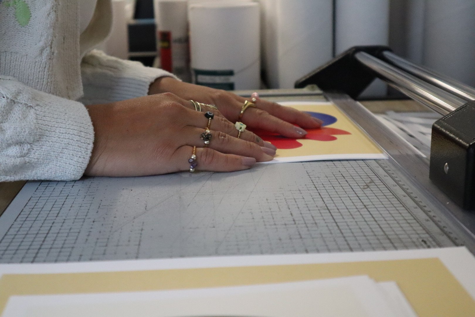 Person wearing rings and a white sweater, working with colorful printed paper on a paper cutter in an art or craft studio.