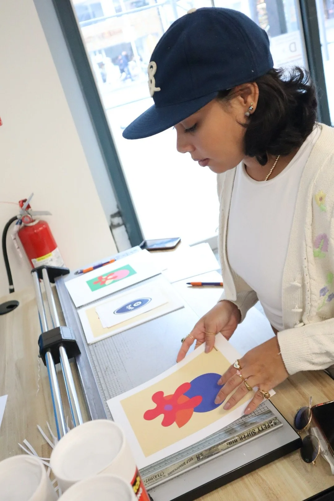 Woman wearing a navy blue cap and white cardigan working on colorful abstract artwork at table with paper, markers, and scissors.