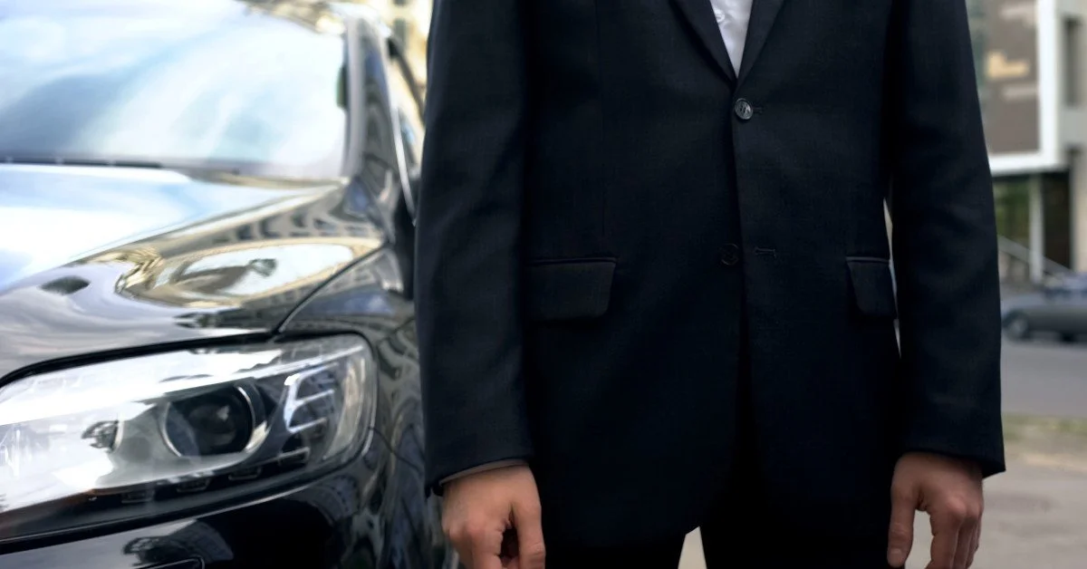 A man wearing a black suit stands next to the front of a black car.