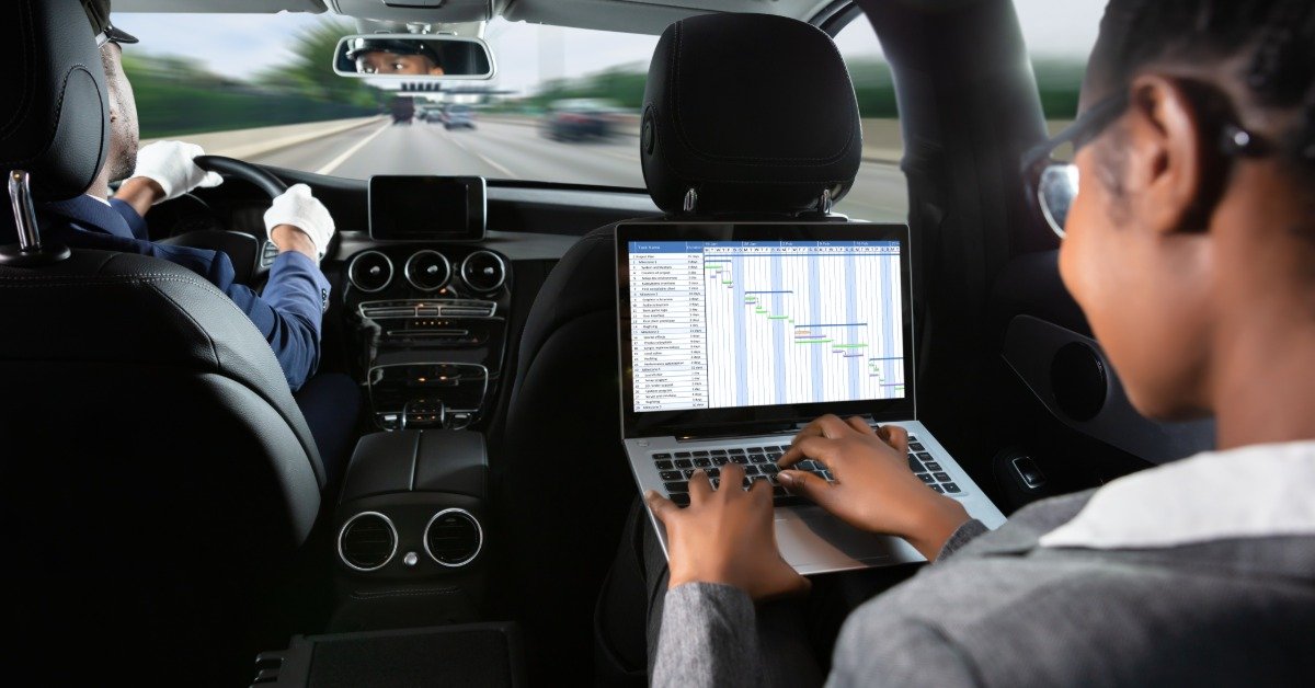 A businesswoman working on a laptop in the back seat of a chauffeured vehicle while a driver navigates highway traffic.