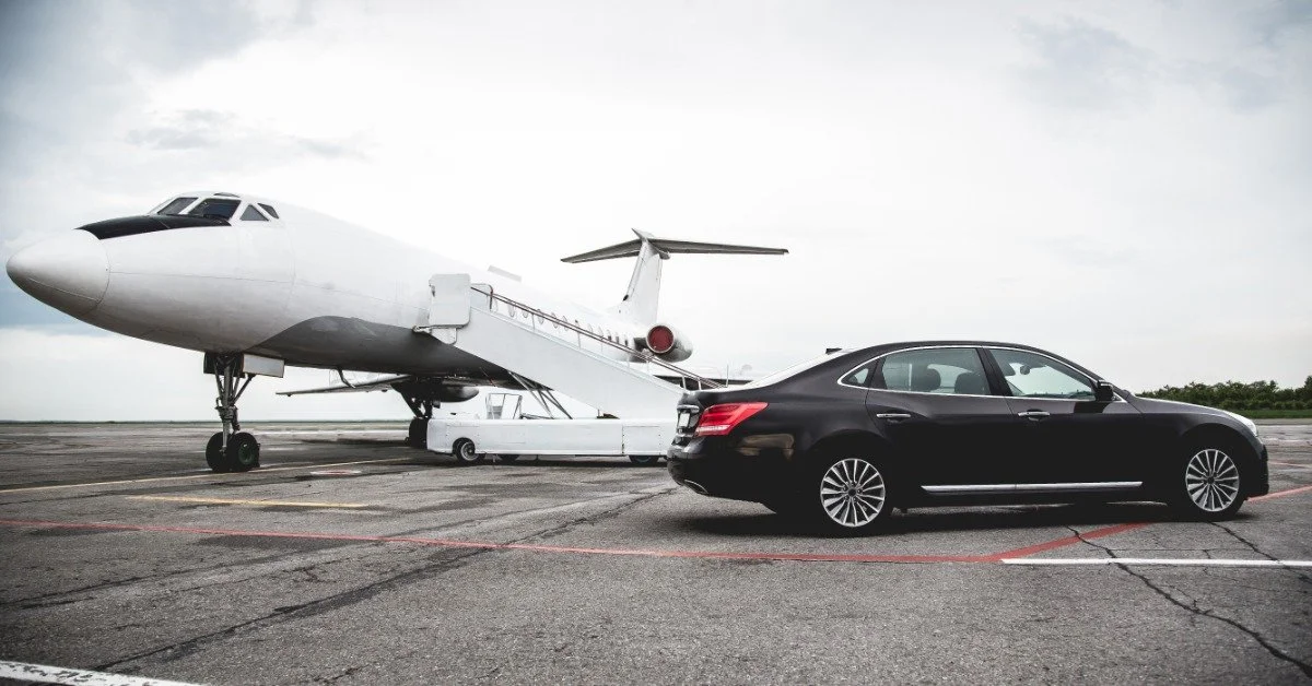 A luxury black sedan parked beside a private jet on an airport tarmac.
