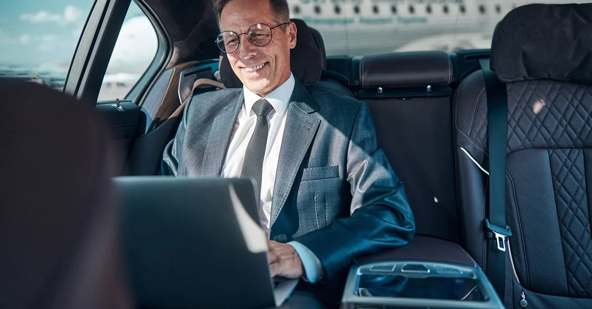 A smiling businessman wearing a gray suit and glasses works on his laptop in the back of a vehicle with a black leather interior.