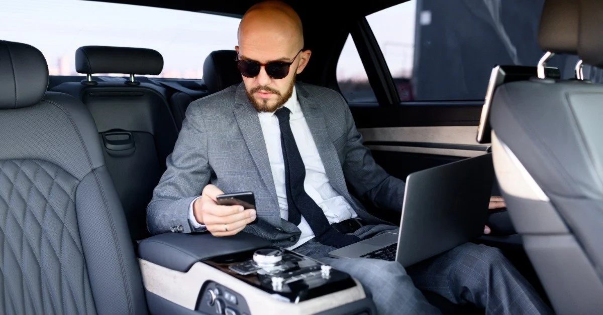 A businessman in a gray suit and sunglasses using a phone and laptop in the back of a luxury vehicle.