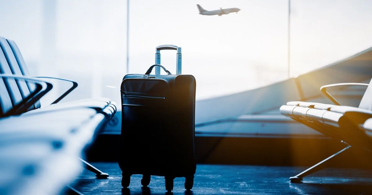 A black suitcase sitting between two rows of seats in an airport terminal, with a large window behind it. A plane takes off in the background.