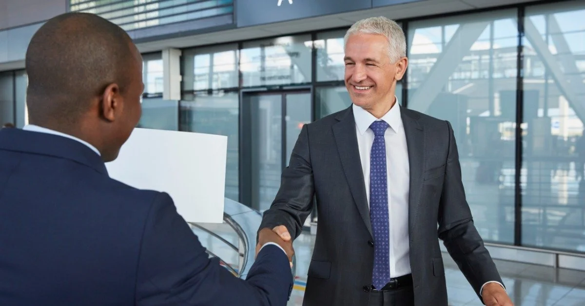 A chauffeur wearing a dark suit greeting a business traveler with a handshake in an airport arrivals terminal.