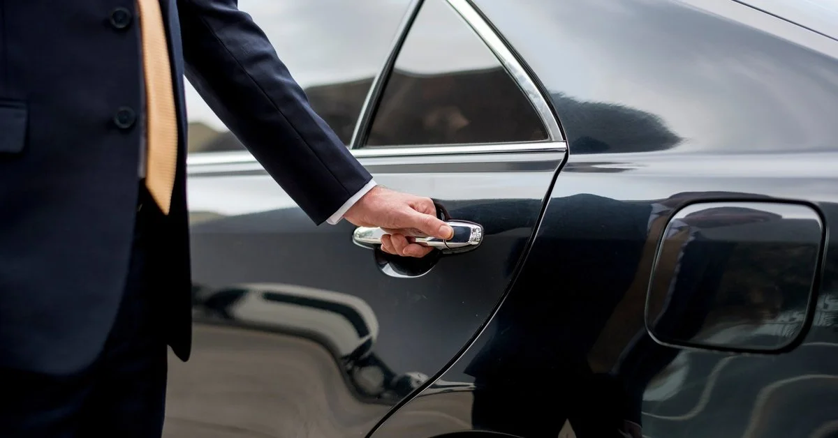 A chauffeur wearing a black suit, placing his hand on the handle of a black car.