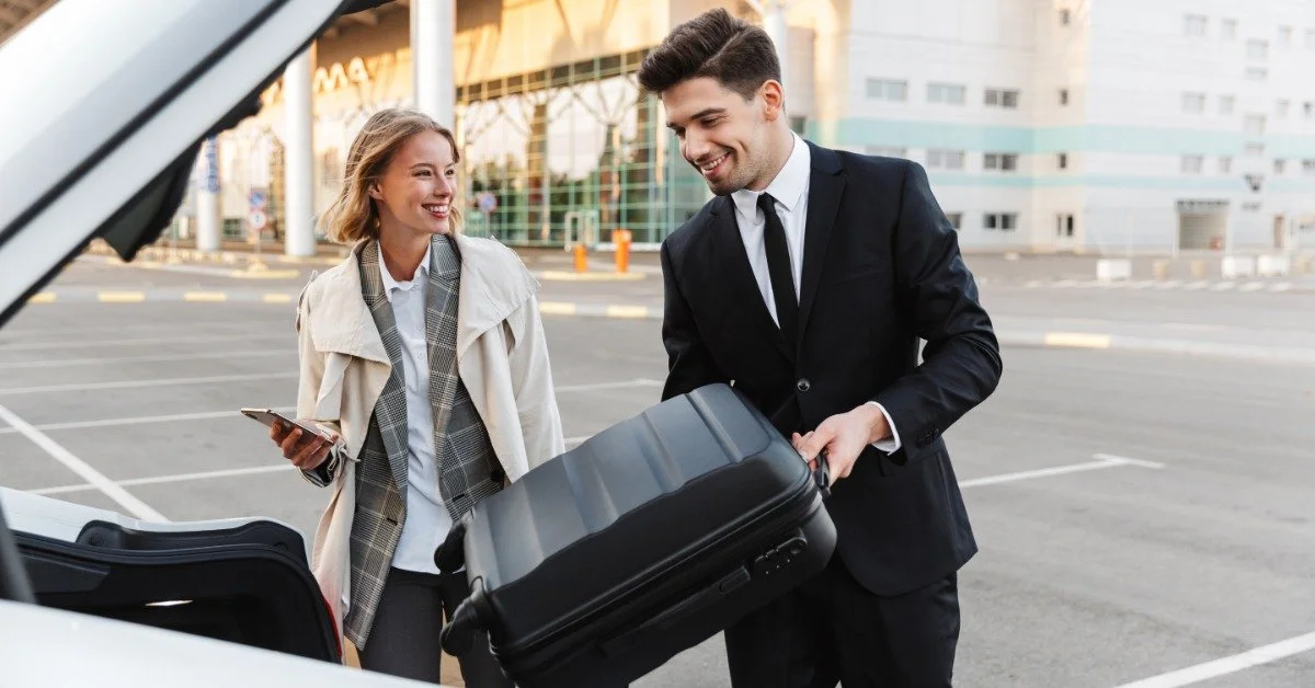 A professional chauffeur loading a passenger’s suitcase into a vehicle outside an airport terminal, with the female passenger smiling and looking on.