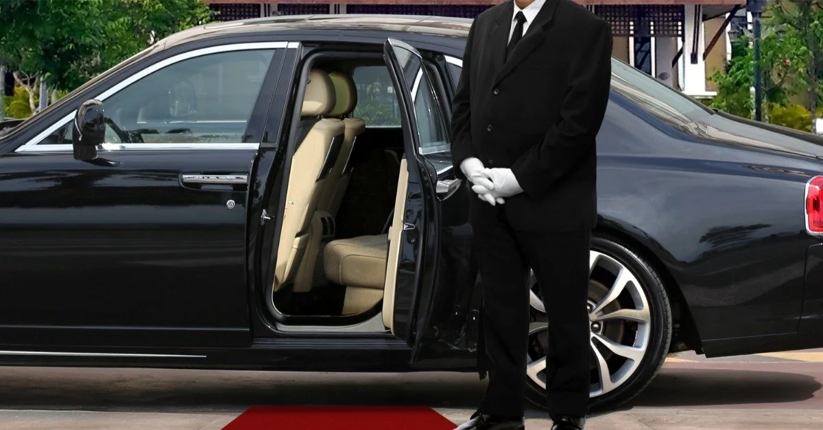 A professional chauffeur in a black suit and white gloves stands beside a black sedan with its back door open, a red carpet leading to the vehicle.