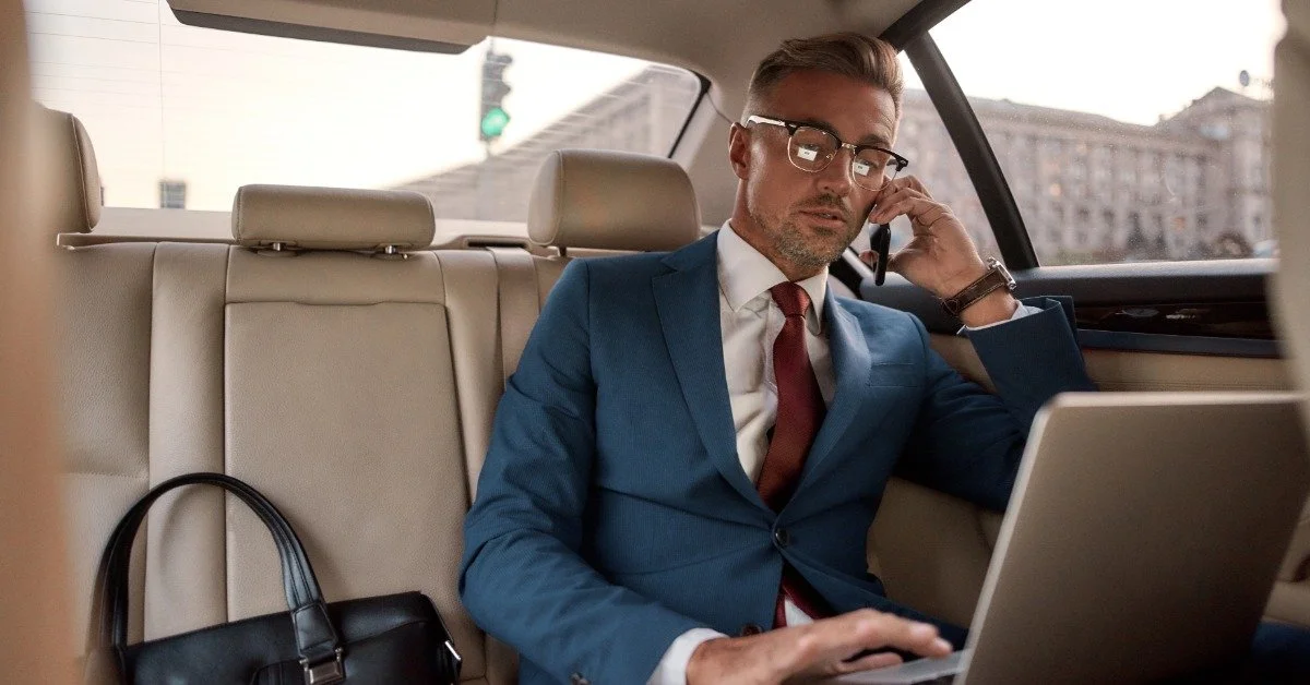 A business executive in a suit working on a laptop and talking on a phone in the back seat of a luxury car.