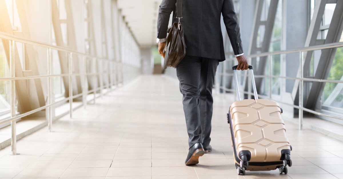 A businessman in a black suit walking through an airport corridor, pulling a white rolling suitcase and carrying a briefcase.
