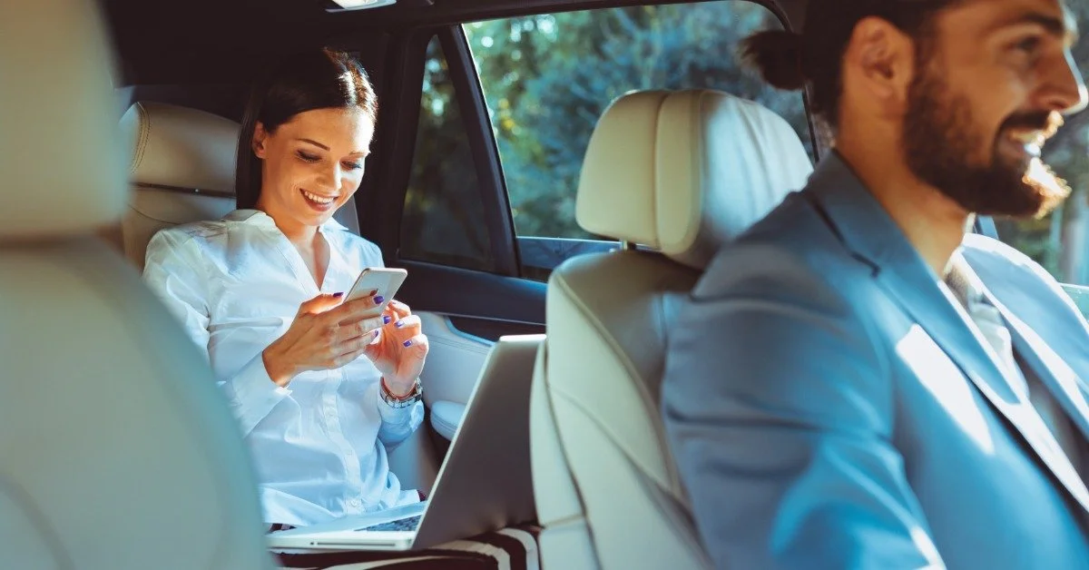 A woman business traveler relaxing in the back seat of a luxury vehicle while using a smartphone and laptop as a chauffeur drives.