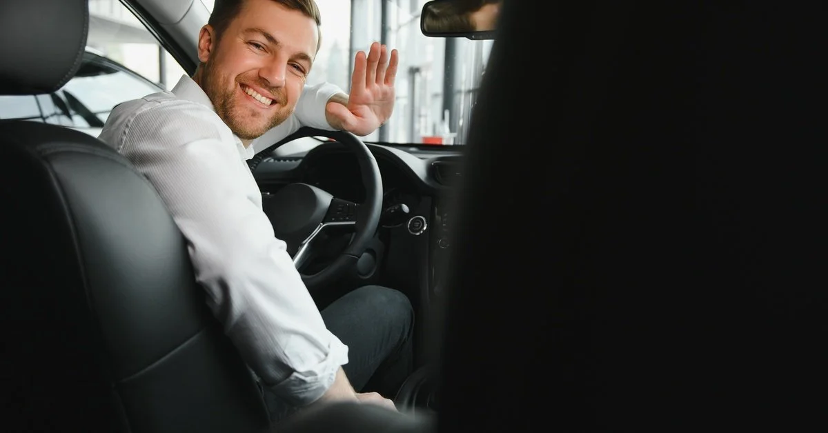 A professionally dressed man smiles and waves from the front seat of a vehicle with a black leather interior.