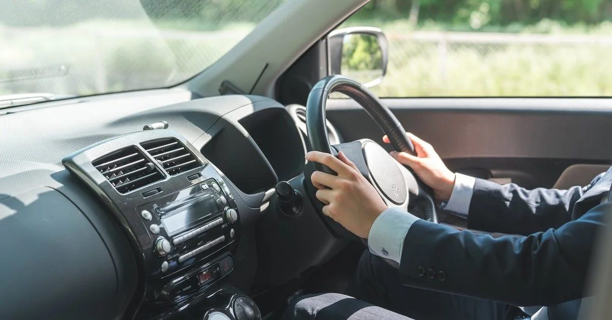 A man wearing a black suit grips the steering wheel of a vehicle with a black interior.