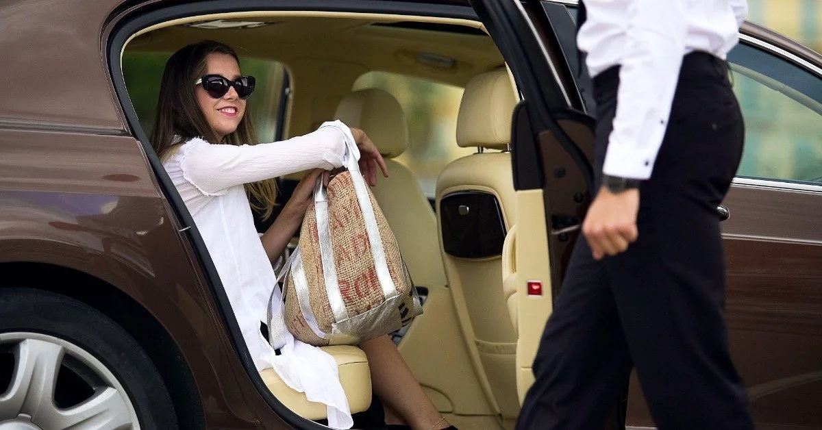 A chauffeur holds open a luxury sedan door for a smiling female passenger seated in the back.