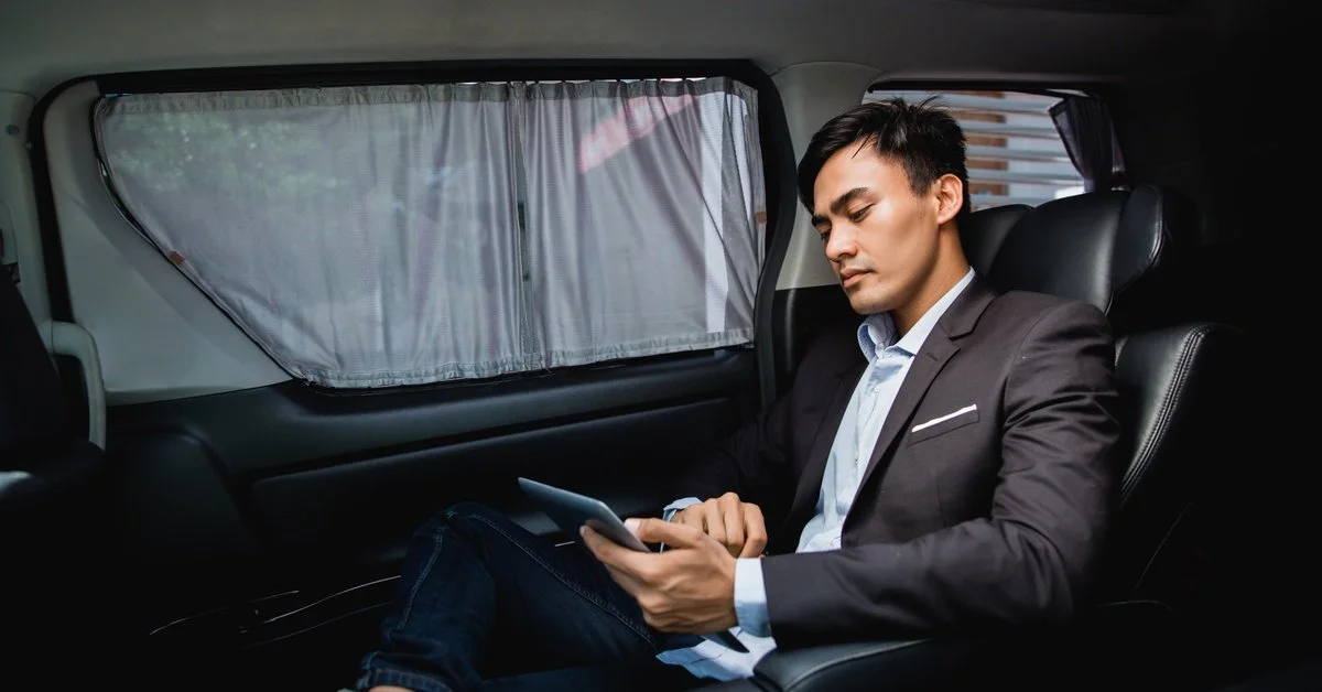 A businessman looks at his tablet as he sits in the back seat of a vehicle with a black leather interior and curtained windows.