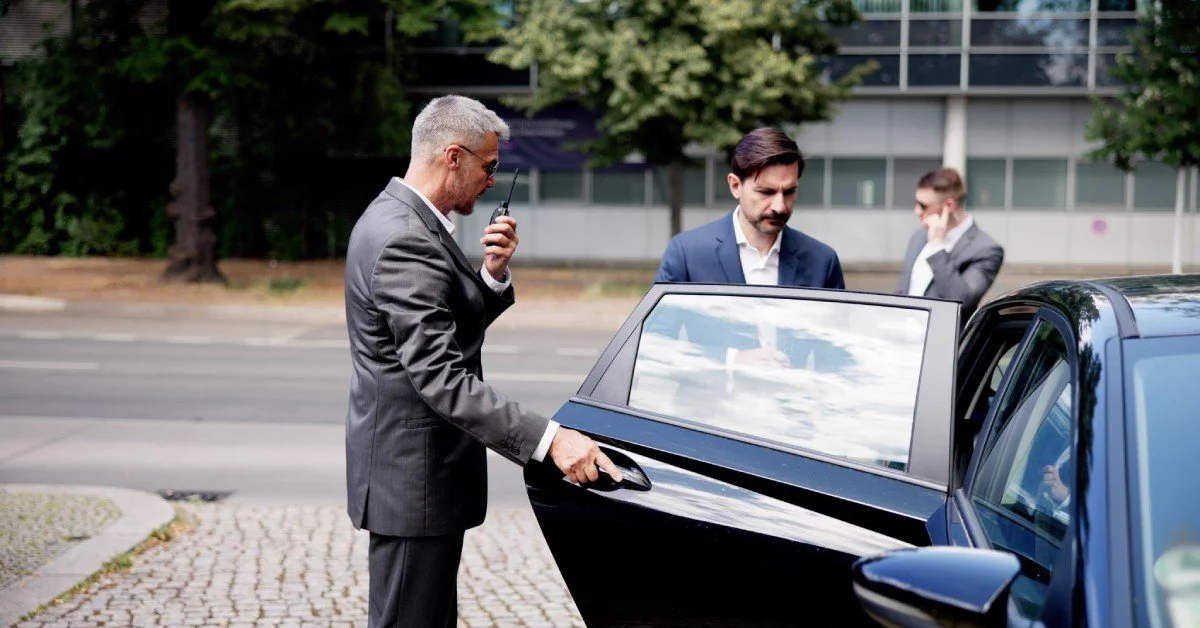 A man speaking into a walkie-talkie opens the door of a black car for another man wearing a business suit.