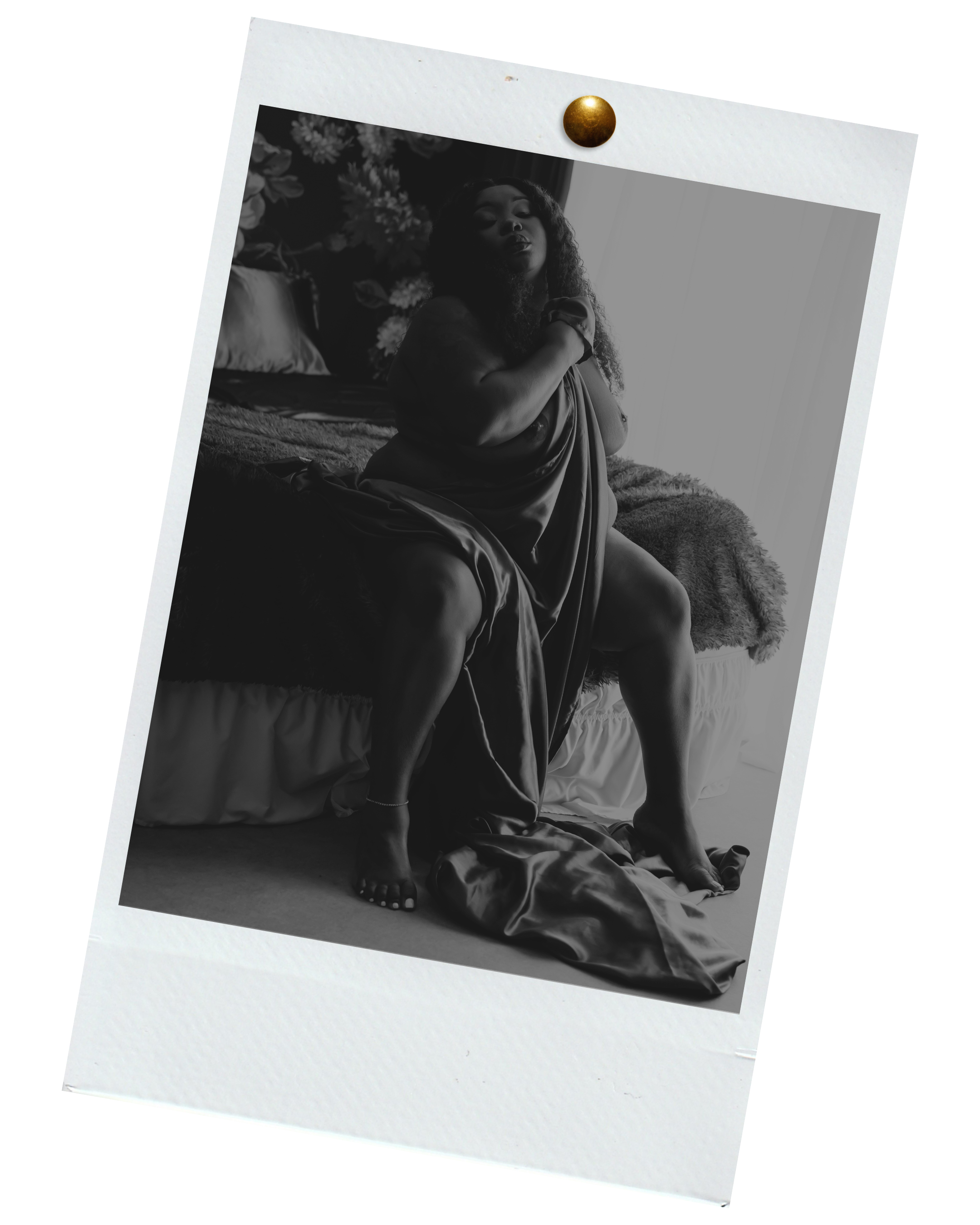 A black and white photo of a woman sitting on a bed, draped in satin fabric, with one arm across her chest and the other resting on her knee, in a softly lit room.