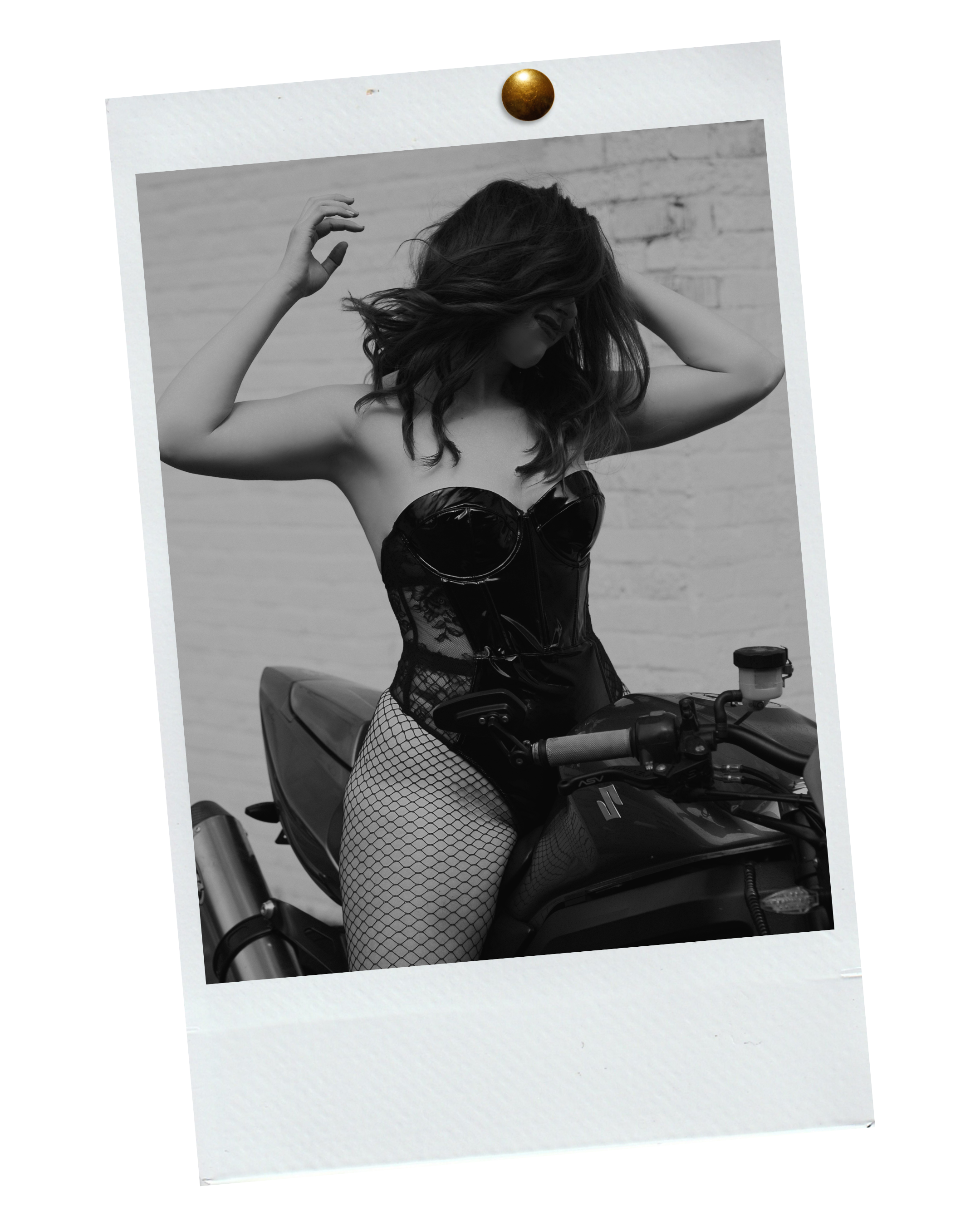 Black and white photograph of a woman with wavy hair, wearing a shiny latex corset, fishnet stockings, and posing on a motorcycle with a background of a brick wall. The image has a Polaroid-style border with a gold push pin at the top.