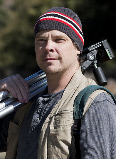 A man wearing a knit beanie hat, carrying a tripod on his shoulder, and wearing a backpack, outdoors with trees in the background.