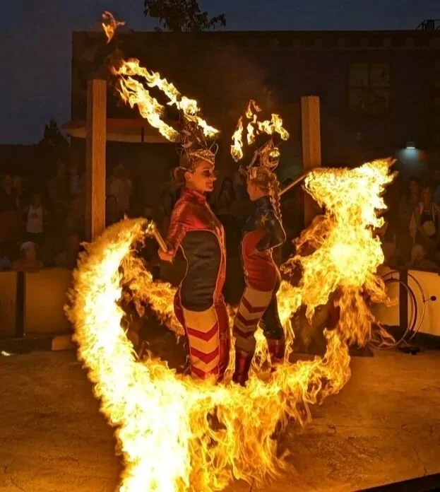 Two performers in elaborate costumes perform a fiery dance surrounded by flames at night.